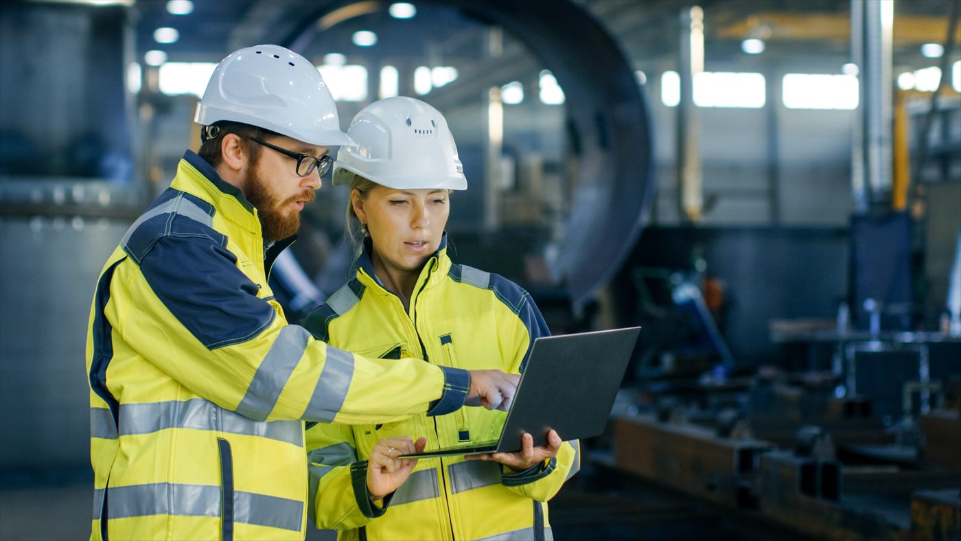 Dos técnicos con casco y chaleco de seguridad revisan datos en un portátil dentro de una planta industrial